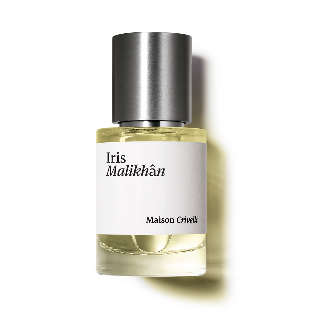 A bottle of Iris Malikhân by Maison Crivelli, featuring a cylindrical silver cap and a clear glass body with a white label displaying the name, embodies an addictive accord with hints of powdery gourmand leather.