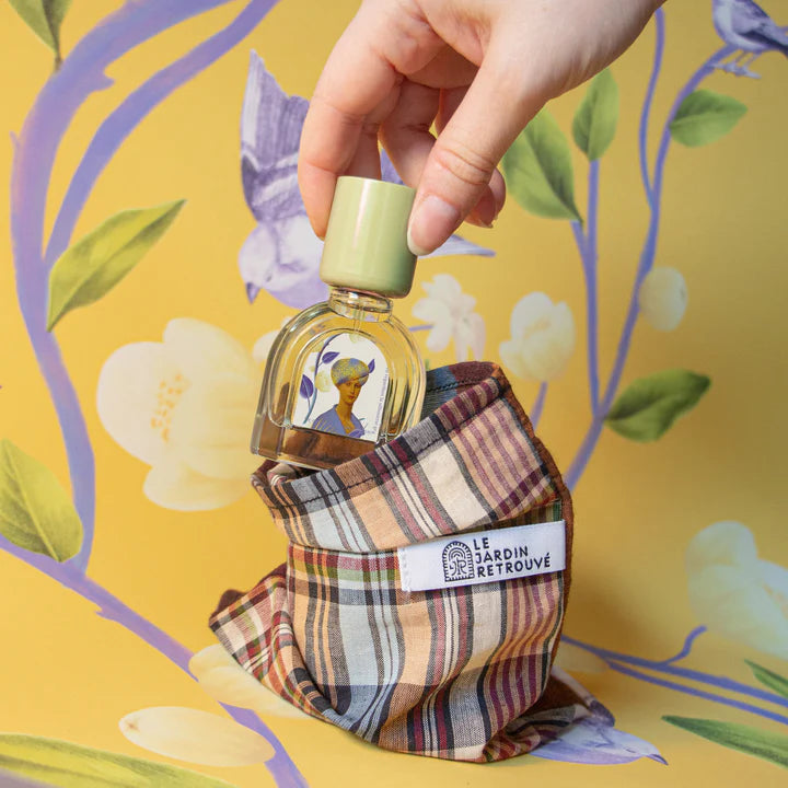 A hand holds a Jasmin Majorelle bottle with a leafy illustration, placing it into a plaid fabric pouch labeled "Le Jardin Retrouvé." The background features a yellow floral and bird design, evoking the essence of Jasmin Majorelle.