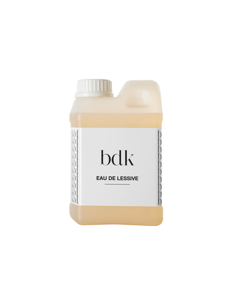 A white plastic container labeled "EAU DE LESSIVE" by BDK Parfums, filled with a pale yellow liquid, featuring a screw-top lid and a side handle for easy pouring. The liquid boasts woody and musky notes, making it suitable for all skin types.