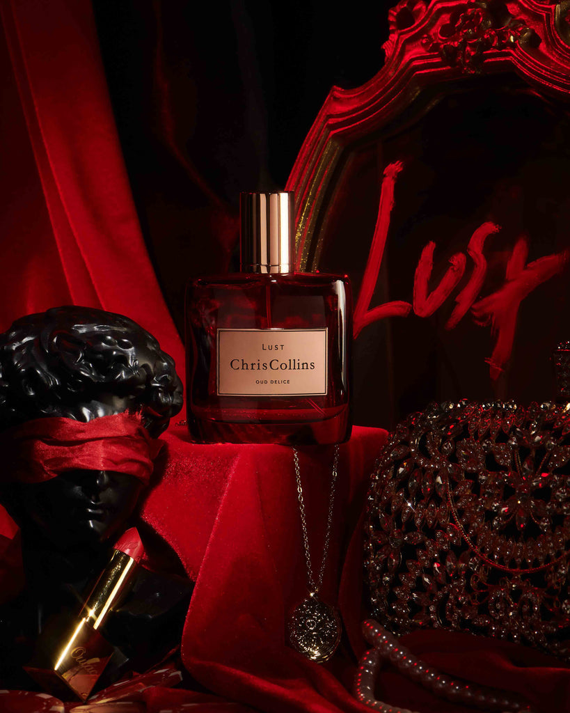 A bottle of Chris Collins LUST | Oud Delice fragrance sits on a red velvet backdrop, accompanied by a black bust with a red blindfold, a gold chain, and an ornate clutch. The scene is heightened with the allure of aphrodisiac perfume and sensual musks that captivate the senses.