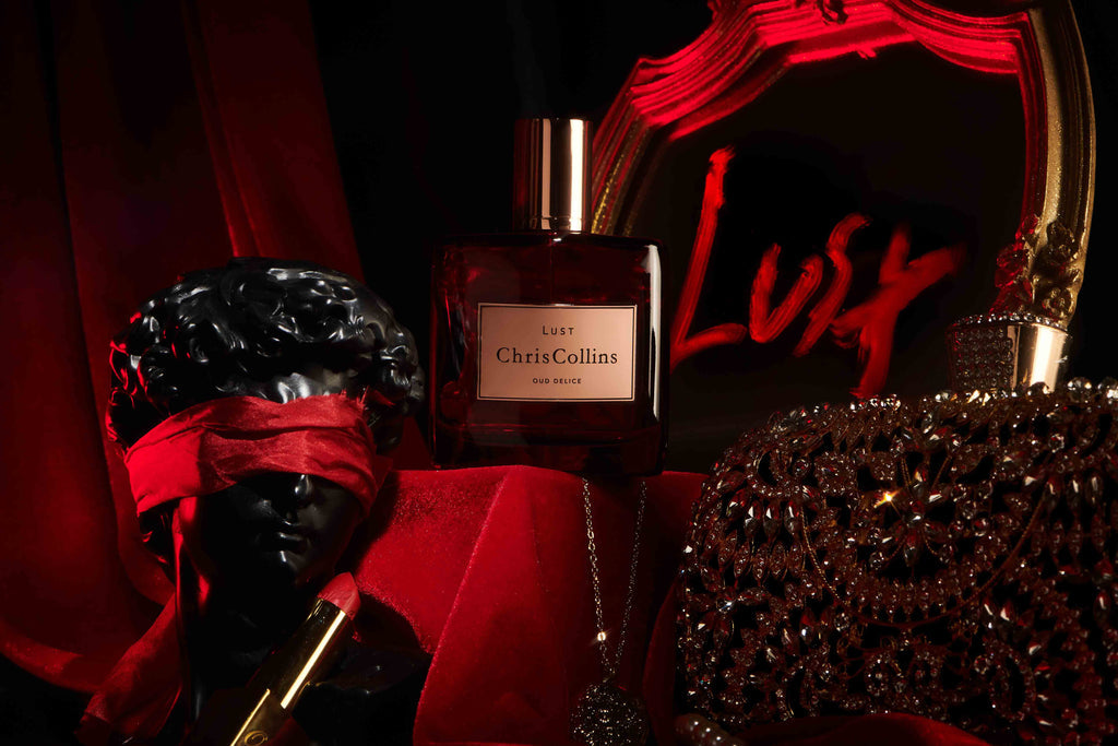 A perfume bottle labeled "Chris Collins LUST | Oud Delice" is displayed on red fabric, next to a blindfolded black statue and a jeweled object, with the word "Lust" written in red on a mirror in the background. The scene evokes the allure of an aphrodisiac perfume, hinting at sensual musks within.