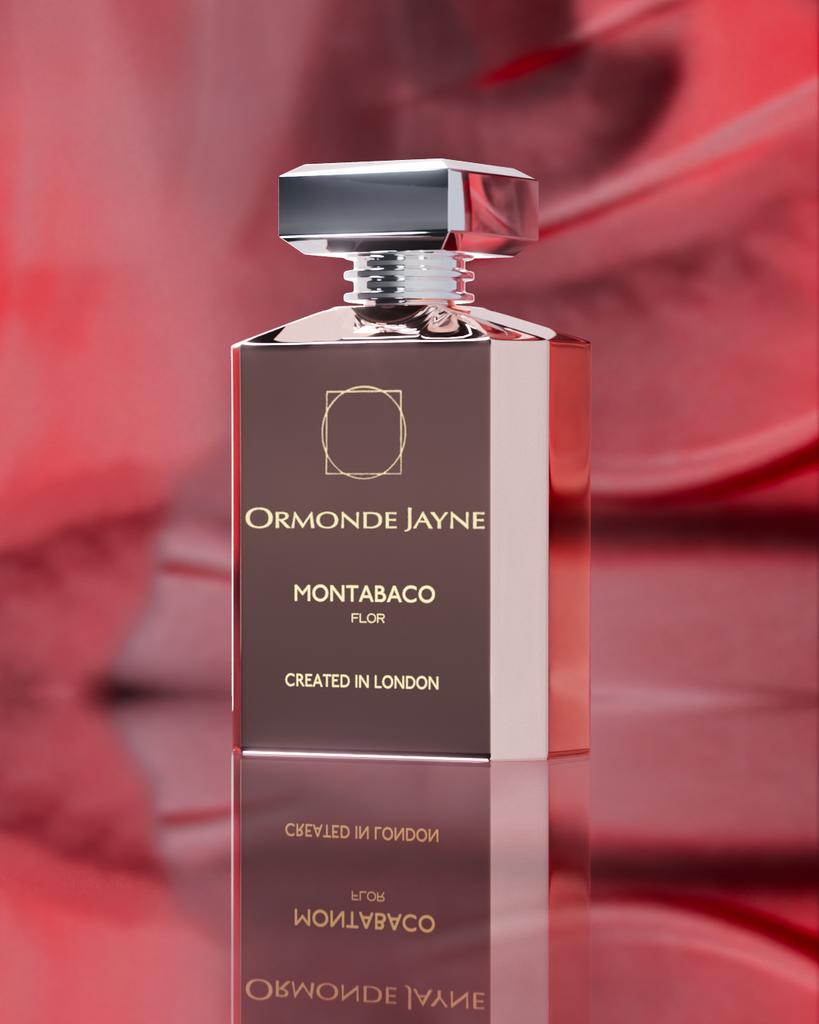 A bottle of Montabaco Flor by Ormonde Jayne with a reflective surface set against a red and pink blurred background. The rectangular bottle, featuring a metallic cap and printed label, exudes elegance with its sandalwood base underpinning the rose jasmine blend of this floral perfume.