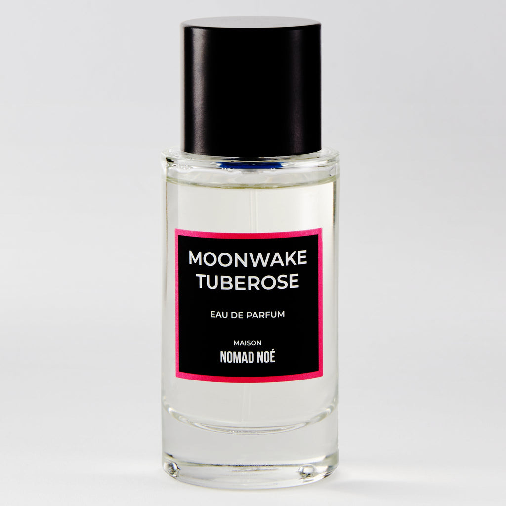 A clear glass bottle of "Moonwake Tuberose" eau de parfum by Nomad Noe, adorned with a black cap and a pink-bordered label, boasts notes of Tuberose Absolute and Sandalwood.