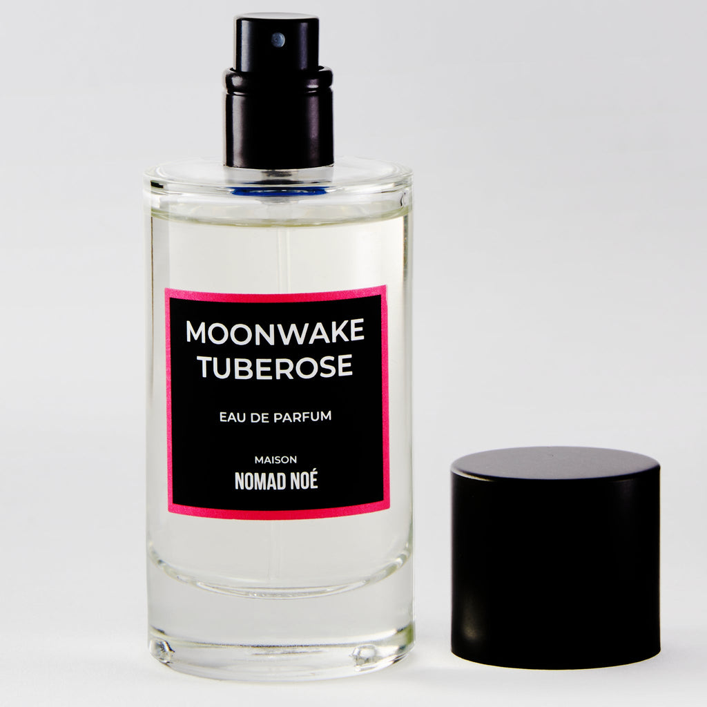 A clear glass bottle labeled "Moonwake Tuberose" by Nomad Noe, infused with Tuberose Absolute and warm hints of Vanilla, stands elegantly with a black cap placed beside it.
