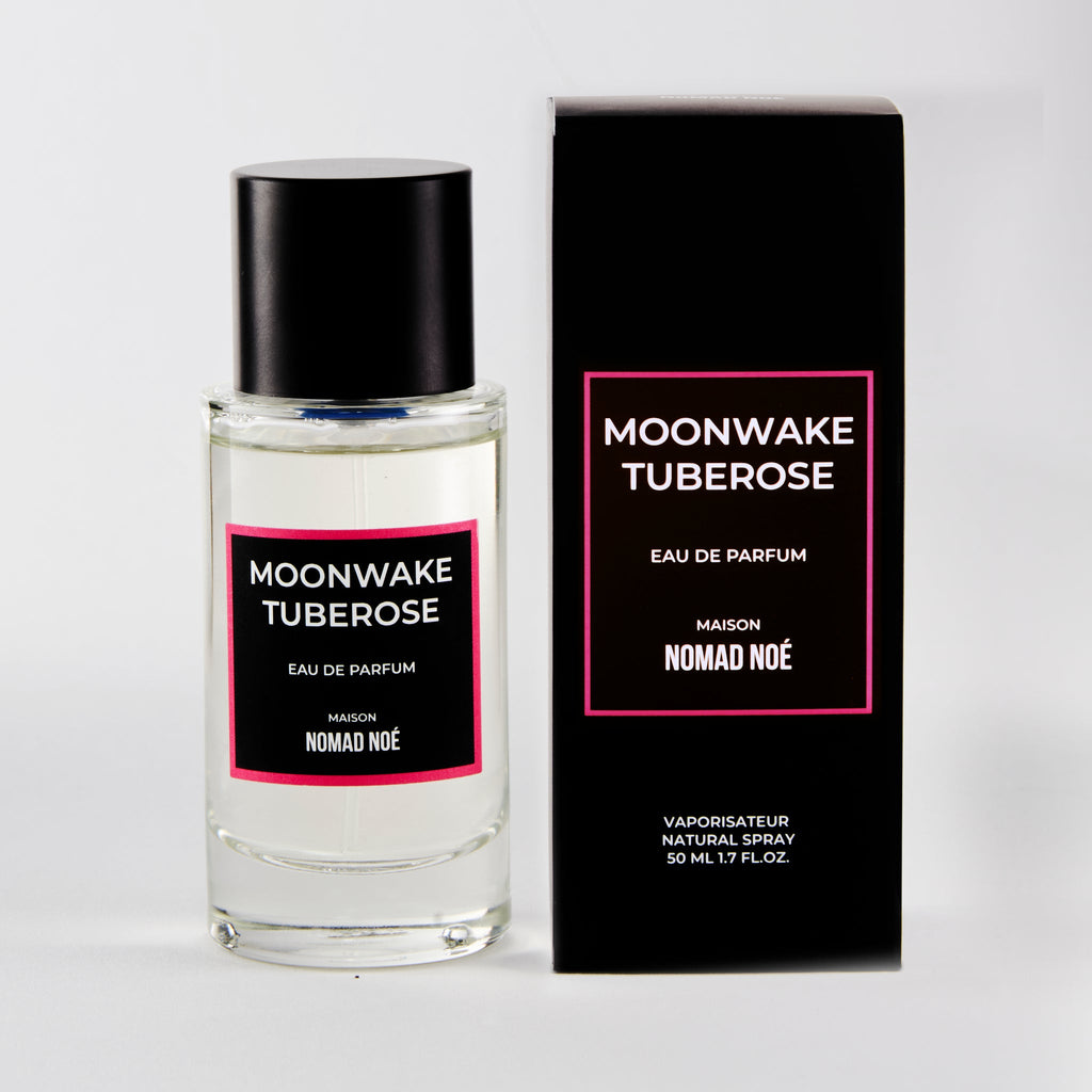 A bottle of "Moonwake Tuberose" eau de parfum by Nomad Noe stands beside its black and pink box. The 50 ml (1.7 fl oz) bottle contains a captivating blend of Tuberose Absolute and Sandalwood, creating an irresistible fragrance.