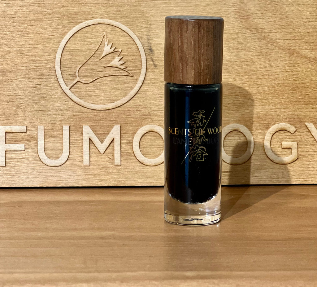 A small bottle of Plum In Cognac perfume by Scents of Wood, with a wooden cap, stands on a wooden surface in front of a wooden backdrop inscribed with "FUMOLOGY" and adorned with a flower logo, exuding subtle hints of vanilla.