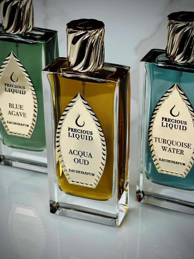 Three perfume bottles labeled "Blue Agave," "Acqua Oud," and "Turquoise Water" by Precious Liquid, with gold and black decorative caps and labels, are arranged in a row on a reflective surface reminiscent of the calm beauty of Caribbean waters. Each fragrance promises an exotic escape.