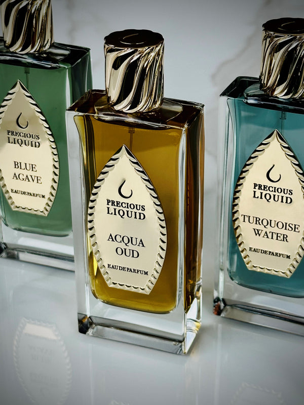 Three perfume bottles labeled "Blue Agave," "Acqua Oud Limited Edition," and "Turquoise Water" are displayed in a row. Each bottle has a distinctive liquid color, a gold label with a leaf design, and promises woody freshness with Acqua Oud Limited Edition by Precious Liquid delivering a long-lasting trail.