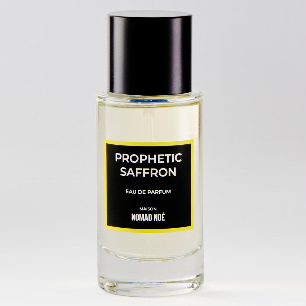 A clear glass bottle of Nomad Noe's "Prophetic Saffron" Eau de Parfum with a black cap and label on a white background, featuring the warm allure of saffron essence intertwined with woody/spicy notes.