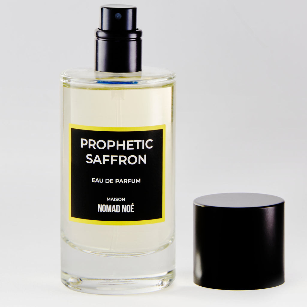 A clear glass bottle of "Prophetic Saffron" eau de parfum by Nomad Noe, infused with saffron essence, with a black spray cap removed and placed nearby.