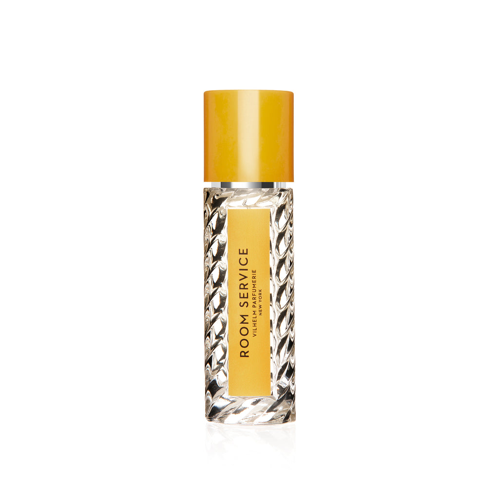 A clear glass bottle with a yellow cap, containing a perfume labeled "Room Service" by Vilhelm Parfumerie on a yellow and white label, reminiscent of flower petals.