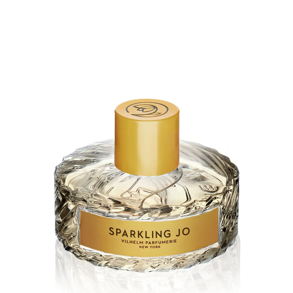 A clear, faceted glass bottle of Vilhelm Parfumerie's "Sparkling Jo" perfume, featuring a gold cylindrical cap and a matching gold label on the front, crafted by the master perfumer in New York.