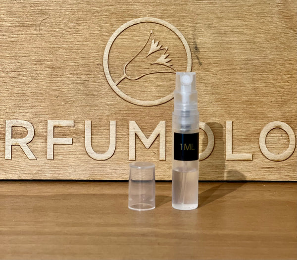 A small 1ml spray bottle of Nicolaï's Oud Sublime Elixir, containing transparent liquid, rests on a wooden surface. Behind it stands a wooden board engraved with "PERFUMOLOGY" and adorned with a floral logo. Infused with the exotic essence of Cambodian oud, this bottle captures an alluring aroma in every spritz.