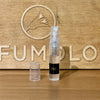 A 2ml spray bottle of Another Kiss by the Fireside by 4160Tuesdays with a black label stands with its clear cap off, set against a wooden background with an engraved bird logo and the word "PERFUMOLOGY." This elegant bottle exudes sophistication.