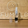 A 2ml spray bottle of Captured by Candlelight from 4160Tuesdays, adorned with a clear cap, is placed in front of a wooden background featuring an engraved bird and the partial word "PERFUMOLOGY," evoking an essence of gourmand woods.