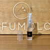 A 2 ml transparent spray bottle with a black label stands on a wooden surface. The cap is placed next to it. A wooden background displays a bird logo and partially visible text, "PERFUMOLO." This scene captures the essence of Eight & Bob perfumes, reminiscent of magical nights of Megève envisioned by Albert Fouquet's Champs de Provence.