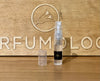 A 2ml clear spray bottle, containing the Nawab of Oudh Intensivo perfume by Ormonde Jayne with its amber and rose blend, features a black label and transparent cap. It is elegantly placed on a wooden surface with a large engraved sign in the background, evoking a sense of emotional complexity.