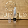 A small, transparent 2ml spray bottle, perfect for dispensing Dragonfly 2021 by Zoologist, with a clear cap is positioned in front of a wooden backdrop featuring the word "PERFUMOLOGY" and a carved bird logo. The scene evokes the tranquility of aquatic florals.