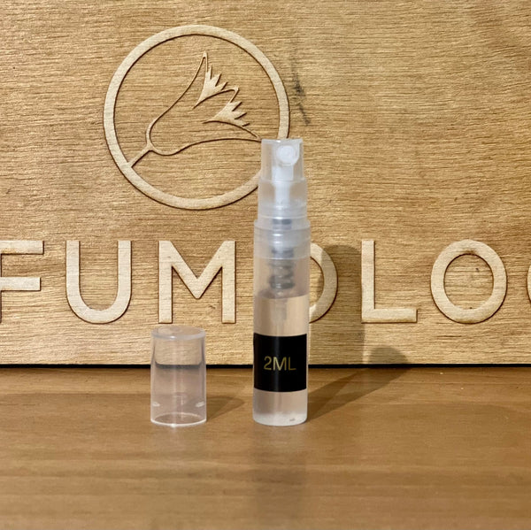 A small, transparent 2ml spray bottle, perfect for dispensing Dragonfly 2021 by Zoologist, with a clear cap is positioned in front of a wooden backdrop featuring the word "PERFUMOLOGY" and a carved bird logo. The scene evokes the tranquility of aquatic florals.