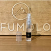 A small 2ml spray bottle of Dark Heart of Old Havana Eau de Parfum from 4160Tuesdays, topped with a clear cap, rests on a wooden surface in front of an engraved wooden sign featuring a bird logo and the partial word "PERFUMOLOG," capturing the essence of Old Havana.