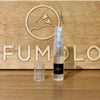 A small, clear 2ml spray bottle filled with Eight & Bob and a removable cap stands in front of a wooden board engraved with a logo and the partial word "PERFUMOLO.