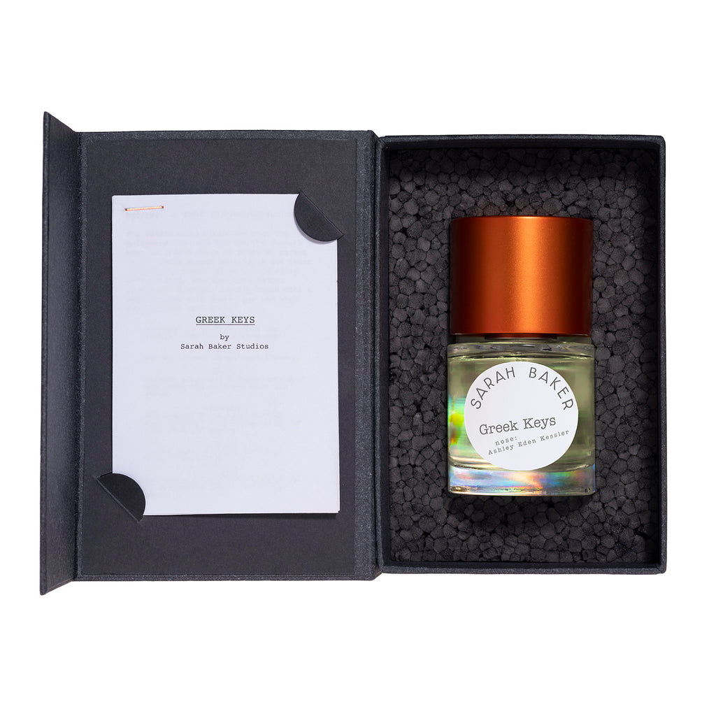 A bottle of Greek Keys by Sarah Baker, adorned with an orange cap and presented in a sleek black box along with a card, exudes a touch of citrus freshness.