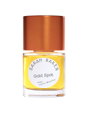 The elegant bottle of Sarah Baker Gold Spot Extrait de Parfum, adorned with a metallic bronze cap and a white label crediting Chris Maurice as the perfumer, holds a rich yellow liquid that hints at its luxurious caramel-oud composition.