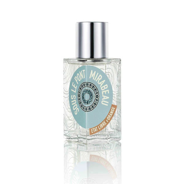 A clear glass bottle of perfume labeled "SOUS LE PONT MIRABEAU" from the brand État Libre d'Orange, featuring a round blue and white emblem and a silver cap, with tantalizing hints of pink pepper and sandalwood.