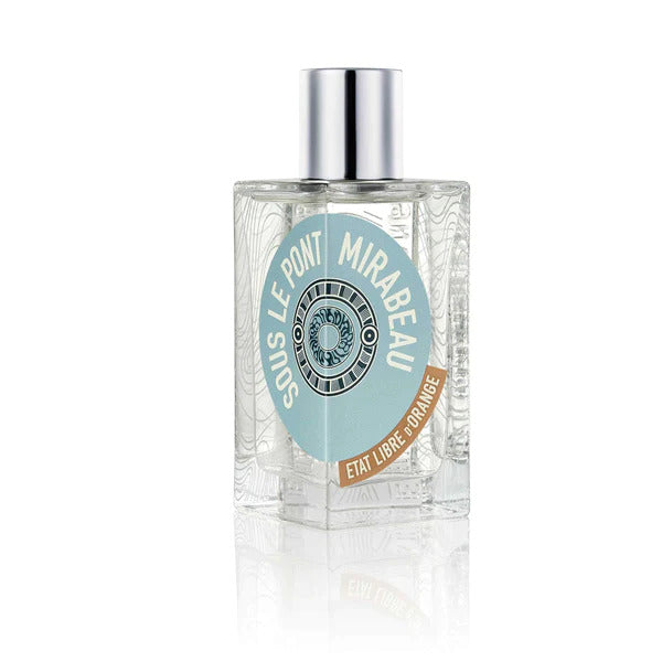 A clear glass bottle of "SOUS LE PONT MIRABEAU" perfume by État Libre d'Orange, with a silver cap and a blue and white circular label on the front, embodies a sophisticated blend of pink pepper and sandalwood notes.