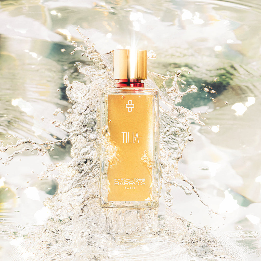 A bottle of Marc-Antoine Barrois TILIA is surrounded by splashing water. The bottle has a golden cap and label with a minimalist design, embodying elegant floral scents.