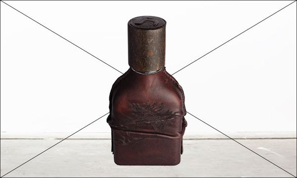 A dark brown, rectangular leather-wrapped flask with an ornate design stands upright on a light-colored surface. Infused with the rich Leather fragrance of Cuoium and crafted by Orto Parisi, the flask features a weathered metal cap.