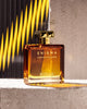 A rectangular bottle of Enigma Pour Homme Parfum Cologne by Roja Parfums, an oriental fragrance with hints of cognac and tobacco, sits on a textured surface with yellow and black diagonal stripes in the background.