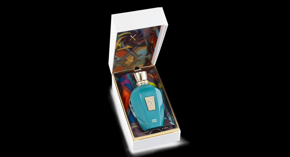 A blue perfume bottle with a gold cap, in a white box lined with colorful, patterned fabric reminiscent of a Mediterranean fruit basket, Erba Pura by Xerjoff.