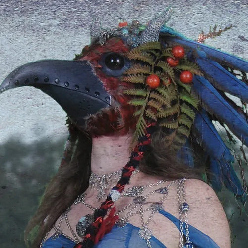 A person wearing an elaborate bird-themed mask with a large beak, adorned with blue feathers, ferns, and red berries reminiscent of the vibrant hues of Ikiryo's Beija Flor, is visible from the side. The background is blurred.