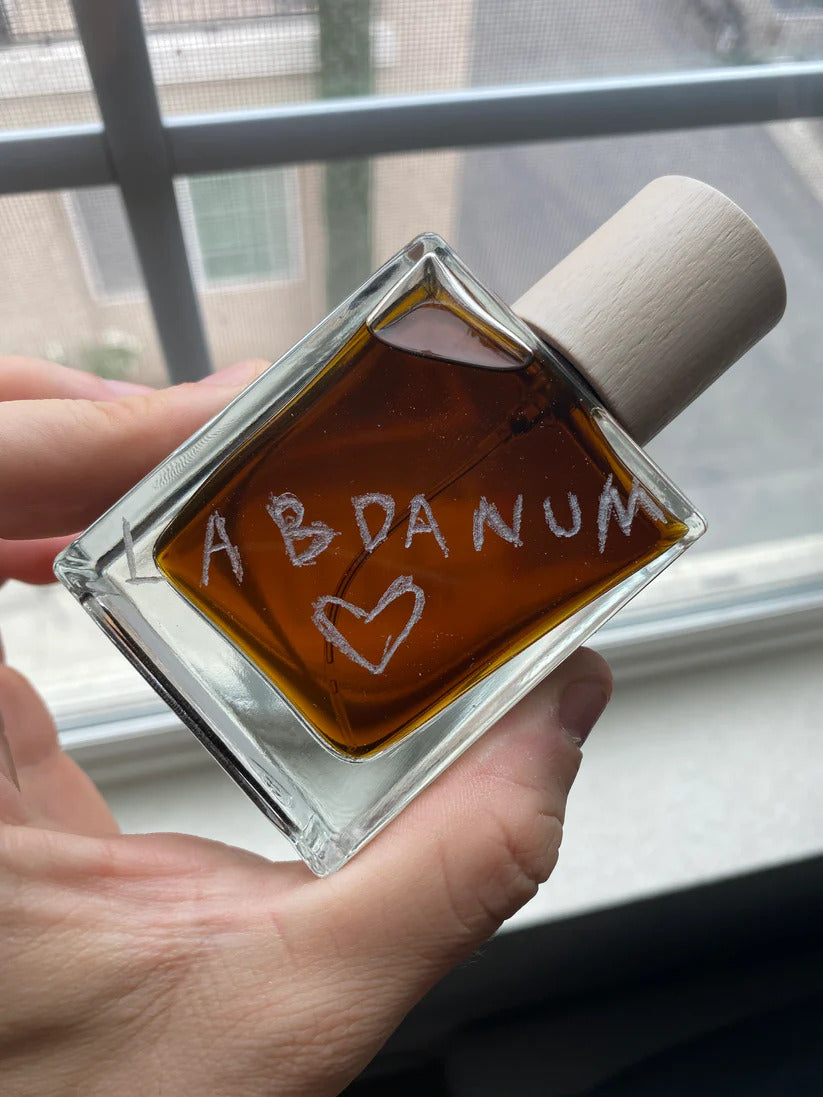 A person holds a square glass perfume bottle with a wooden cap, labeled "Labdanum is my love language" and adorned with a hand-drawn heart, near a window, as warm rays of sunlight highlight the rich amber hue of the fragrance inside.