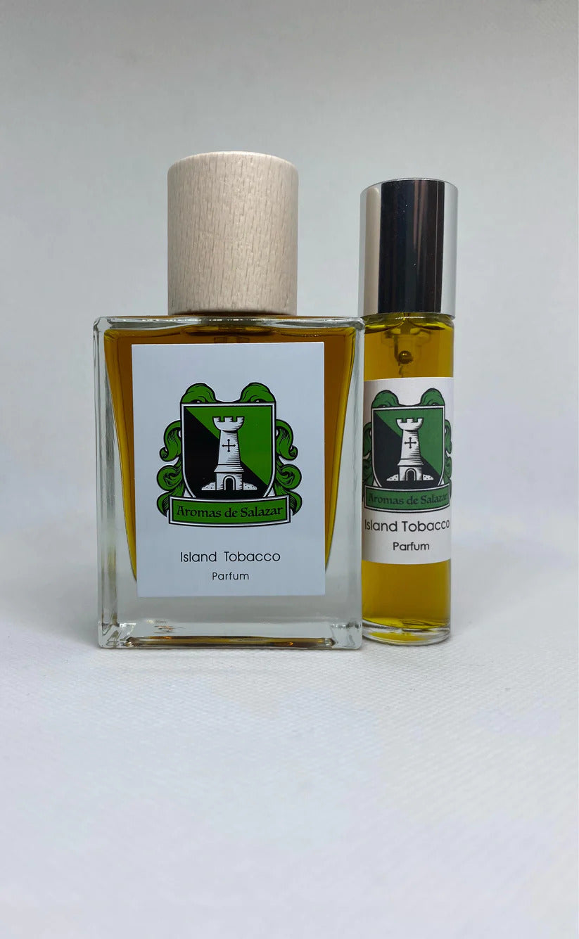 Two perfume bottles, one square and one cylindrical, labeled "Island Tobacco" by Aromas de Salazar. Both feature a green and white logo with a tower and foliage, delivering a rich tobacco aroma blended with a refreshing tropical scent.