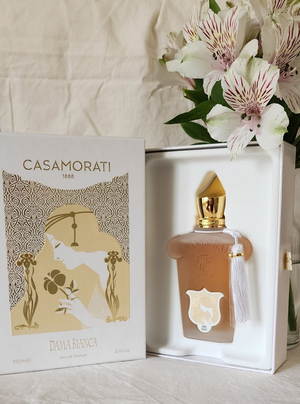 A Casamorati Dama Bianca eau de parfum bottle and its packaging are displayed next to a bouquet of white and pink flowers, evoking a romantic liaison with the elegance of Italian perfumery.