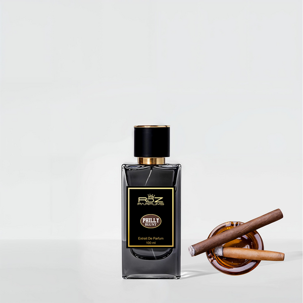 A bottle of "PHILLY Blunt" extrait de parfum by RDZ Parfums with a black and gold label sits next to an open wooden container holding two rolled cigars, evoking a sweet tobacco aroma that resonates with urban culture.