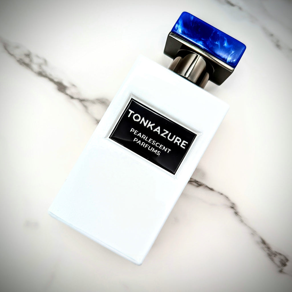 A white perfume bottle labeled "Tonkazure" by Pearlescent Parfums, featuring a blue rectangular cap and set against a marble surface, exudes the sophistication of champagne and the allure of tonka bean.