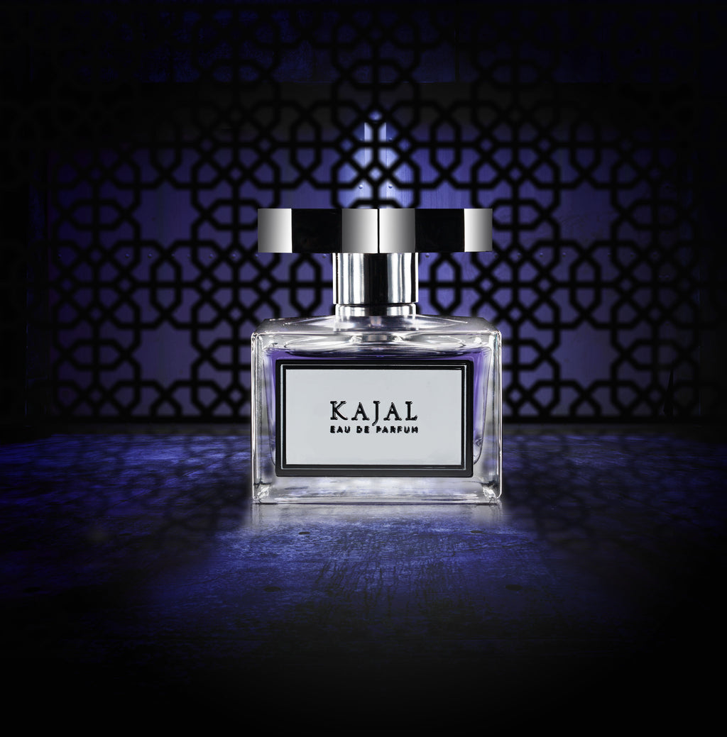 A bottle of Kajal - Classic Collection is centered against a dark, intricately patterned background with a blue hue. The rectangular bottle features a black and silver cap and a label on the front, highlighting the sophisticated fragrance within.