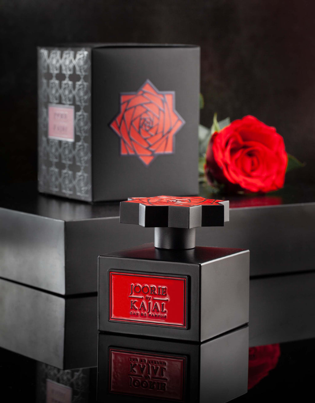 A bottle of Joorie from the Kajal - Wardé Collection, featuring a red rose logo, is elegantly positioned in front of its black packaging that boasts matching red rose graphics, encapsulating the essence of love. A single red rose rests beside the packaging, perfectly complementing the design.