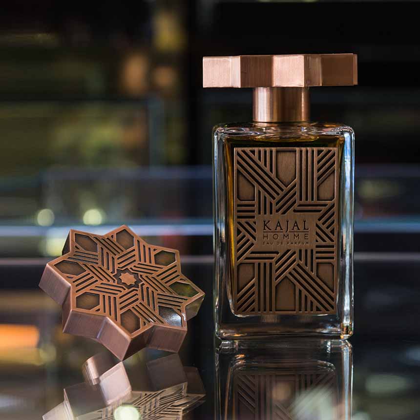 A bottle of Kajal Homme from the classic collection by Kajal Perfumes Paris, adorned with a decorative gold cap beside it on a reflective surface, exudes the sophisticated allure of men’s fragrance.