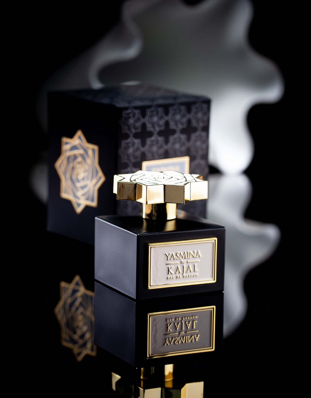 A black and gold perfume bottle labeled "Yasmina" from the Kajal - Wardé Collection, with a scent reminiscent of jasmine flowers, sits in front of an ornate black and gold box against a reflective surface.