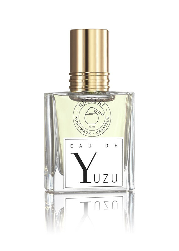 A glass bottle of Nicolaï Eau De Yuzu perfume with a gold cap and black-and-white label, reflecting on a white surface, exudes the refreshing aroma of guaiac wood and patchouli.