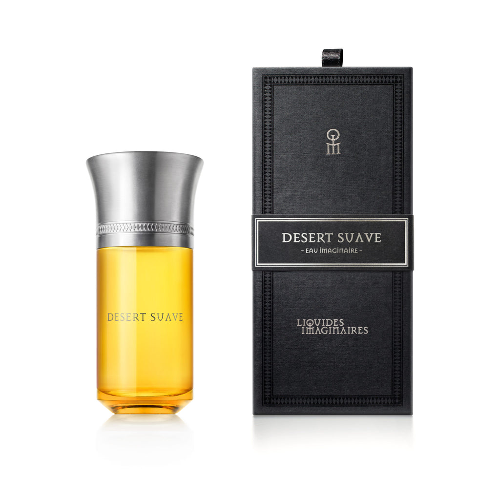 A bottle of Desert Suave perfume with orange-hued liquid and a silver cap is displayed next to its black rectangular box. Infused with notes of cardamom and orange blossom, the fragrance exudes a captivating aura. The box features the product name "Desert Suave" and the brand "Liquides Imaginaires" printed on it.