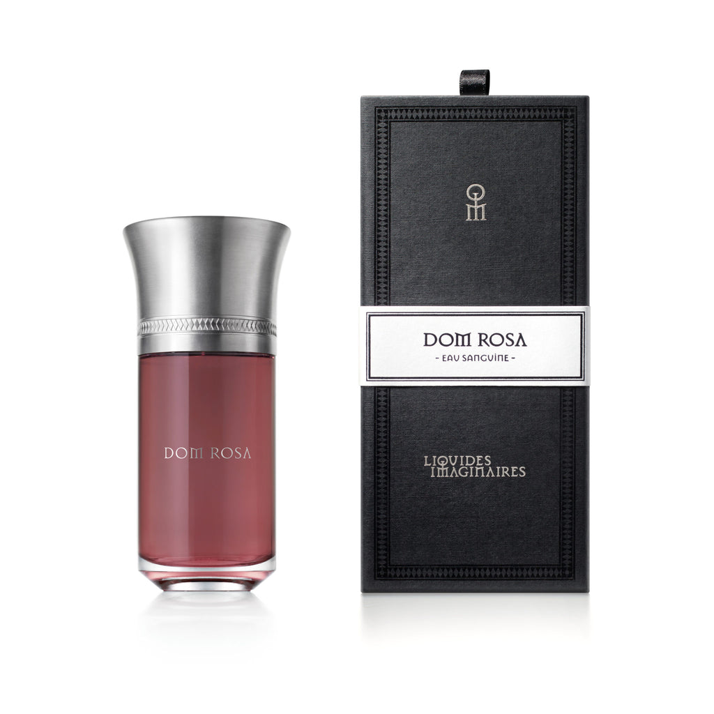 A bottle of Liquides Imaginaires' Dom Rosa Eau Sanguine Amber Floral fragrance with a metallic cap is placed next to its black rectangular box with silver accents, exuding the elegance of Damask Rose and the effervescence of champagne.