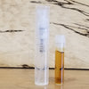 A small transparent spray bottle next to a small cylindrical container filled with a yellow liquid, likely the Sample Subscription Pack from Perfumology, placed on a wooden surface.
