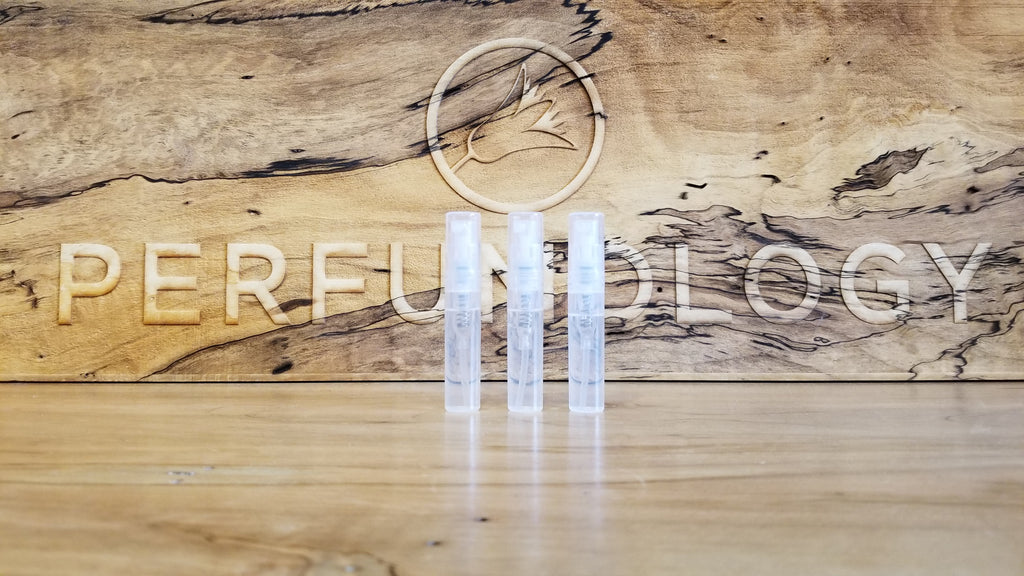 Three small, clear cylindrical containers are placed side by side on a wooden surface with the word "Perfumology" and a bird symbol engraved in the background, showcasing a Sample Subscription Pack that delivers curated perfume samples.
