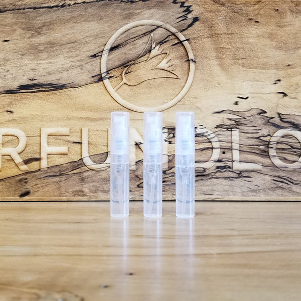 Three small, clear cylindrical containers are placed side by side on a wooden surface with the word "Perfumology" and a bird symbol engraved in the background, showcasing a Sample Subscription Pack that delivers curated perfume samples.