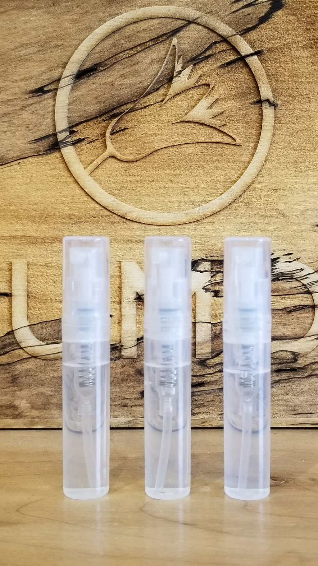 Three clear plastic spray bottles of Perfumology Sample Subscription Pack are arranged in a row in front of a wooden background etched with a bird design and partial text.